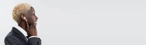 Vista laterale di felice uomo africano americano regolazione auricolare wireless isolato su grigio, banner — Foto stock