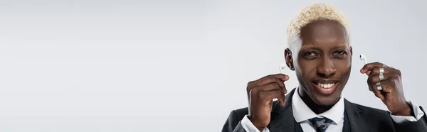 Hombre afroamericano feliz y rubio sosteniendo auriculares inalámbricos aislados en gris, bandera - foto de stock