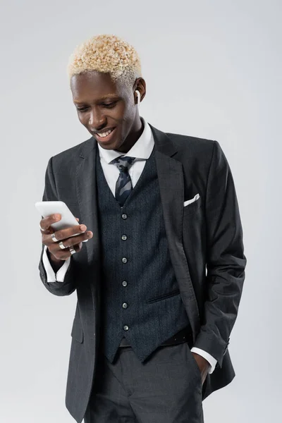 Homem americano africano feliz em fone de ouvido sem fio usando smartphone isolado em cinza — Fotografia de Stock