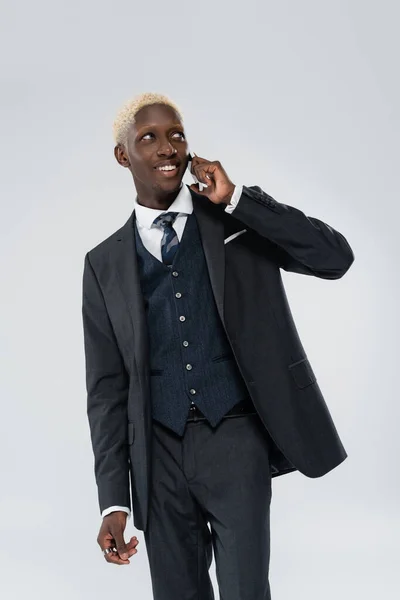Веселий афроамериканський чоловік у костюмі розмовляє на смартфоні ізольовано на сірому — стокове фото