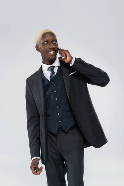 Cheerful african american man in suit talking on smartphone isolated on grey — Stock Photo