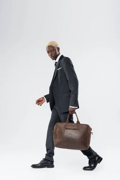 Full length of blonde african american man in suit walking with leather bag on grey — Stock Photo