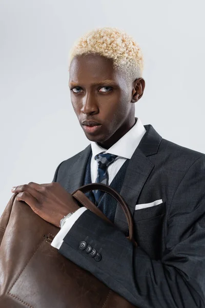 Блондин африканский американец с голубыми глазами, держащий кожаную сумку, изолированную на сером — стоковое фото