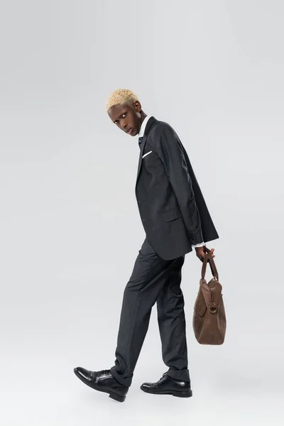 Full length of blonde african american man in suit standing with leather bag on grey — Stock Photo