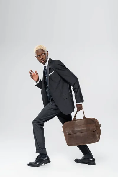 Full length of suspicious african american man in suit walking with leather bag on grey — Stock Photo