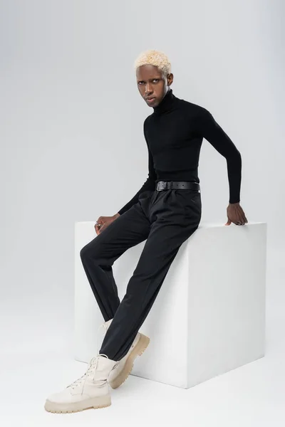 Full length of blonde african american man in turtleneck leaning on white cube on grey — Stock Photo