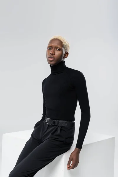 Blonde african american man in turtleneck sitting on white cube isolated on grey — Stock Photo