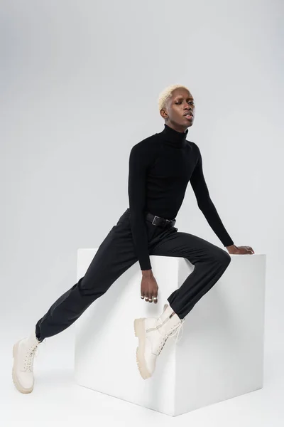 Full length of blonde african american man in turtleneck posing on white cube on grey — Stock Photo