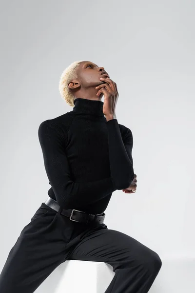 Blonde african american man in turtleneck posing while sitting on white cube isolated on grey — Stock Photo