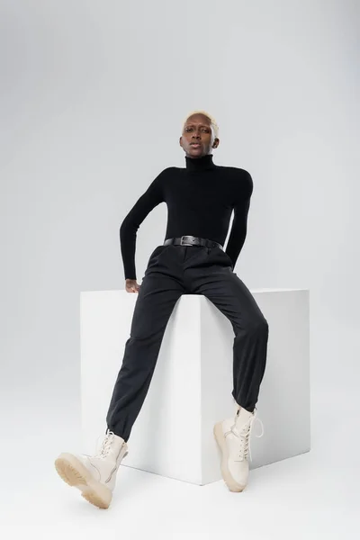 Full length of young african american man in turtleneck posing on white cube on grey — Stock Photo