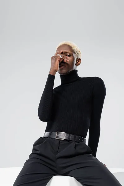 Young african american man in turtleneck touching face while sitting on white cube isolated on grey — Stock Photo