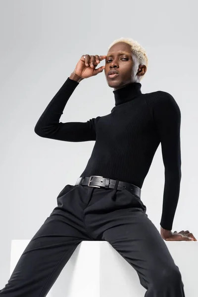 Blonde african american man in turtleneck sitting on white cube and looking at camera isolated on grey — Stock Photo