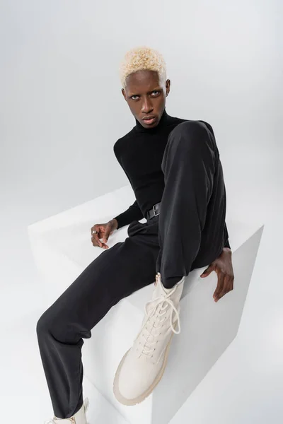 Блондинка афроамериканський молодий чоловік сидить на білому кубі ізольовано на сірому — стокове фото
