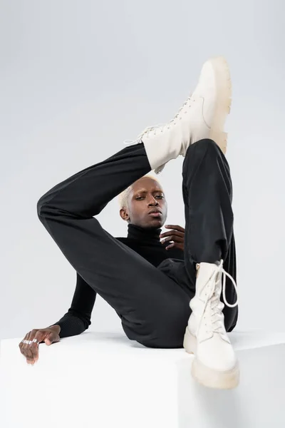 Young african american man in stylish outfit posing on white cube isolated on grey — Stock Photo