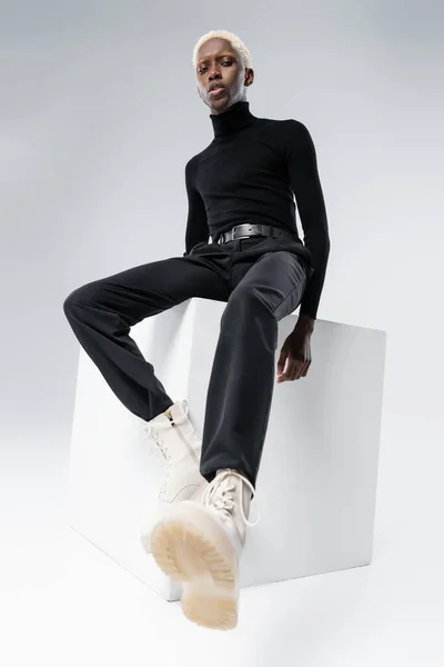 Vista basso angolo di giovane uomo afroamericano in abito elegante seduto sul cubo sul grigio — Foto stock