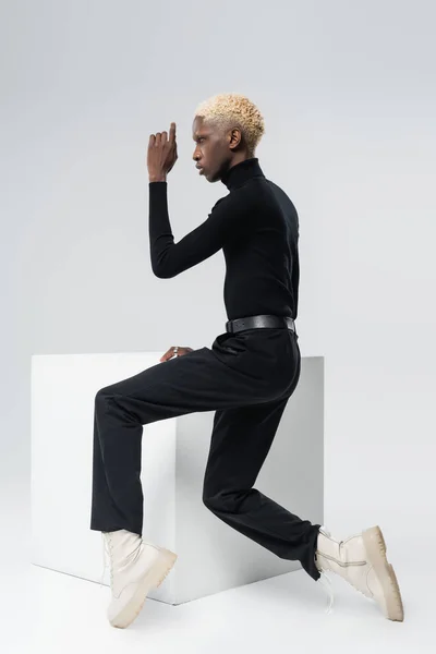 Side view of young african american man in stylish outfit sitting on white cube and posing on grey — Stock Photo