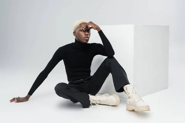 Young african american man in totally black outfit sitting near white cube and posing on grey — Stock Photo