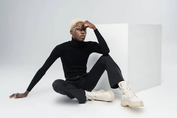 Joven afroamericano en traje totalmente negro sentado cerca del cubo blanco y posando sobre gris - foto de stock