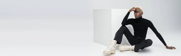 Young african american man in totally black outfit sitting near white cube and posing on grey, banner — Stock Photo