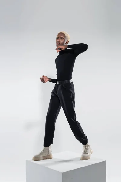 Longitud completa de joven afroamericano hombre en traje totalmente negro de pie en cubo blanco mientras posando en gris - foto de stock