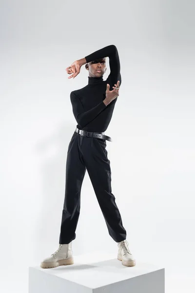 Full length of young african american man in totally black outfit standing on white cube while posing on grey — Stock Photo