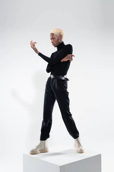 Full length of young african american man in totally black outfit standing on white cube while gesturing on grey — Stock Photo