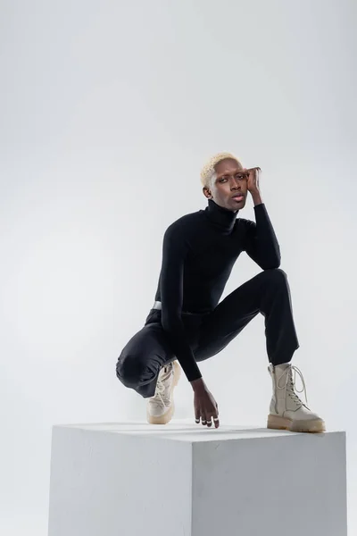 Full length of young african american man in totally black outfit sitting on cube on grey — Stock Photo