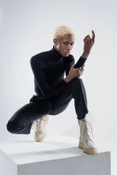 Full length of stylish african american man in totally black outfit sitting on cube on grey — Stock Photo