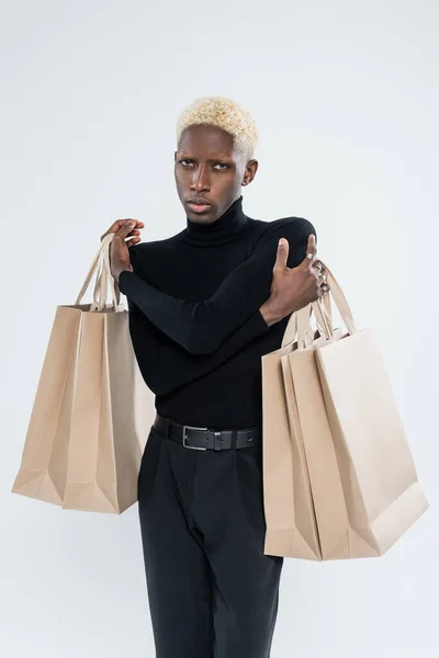 Hombre afroamericano rubio en traje totalmente negro sosteniendo bolsas de compras aisladas en gris - foto de stock