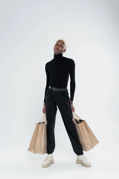 Full length of blonde african american man in totally black outfit holding shopping bags on grey — Stock Photo