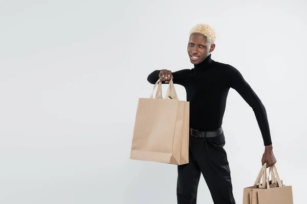 Happy and blonde african american man in totally black outfit holding shopping bags isolated on grey — Stock Photo