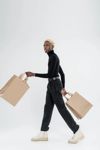 Full length of blonde african american man in totally black outfit walking with shopping bags on grey — Stock Photo