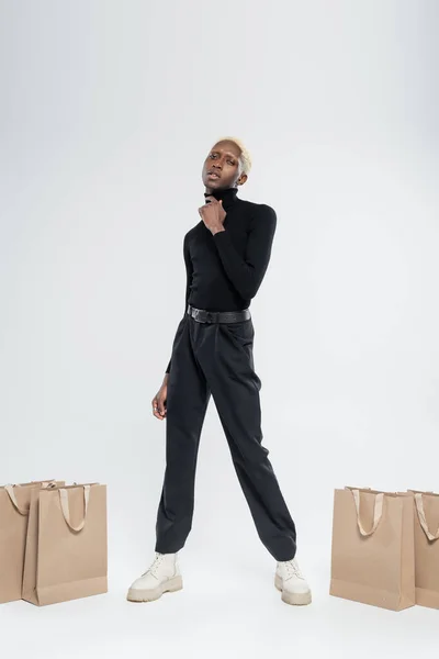 Longitud completa de hombre afroamericano rubia posando cerca de bolsas de papel en gris - foto de stock
