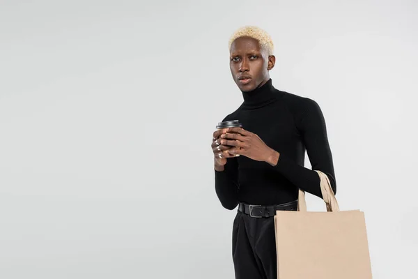 Блондинка афроамериканка стоїть з сумкою для покупок і паперовою чашкою ізольована на сірому — стокове фото
