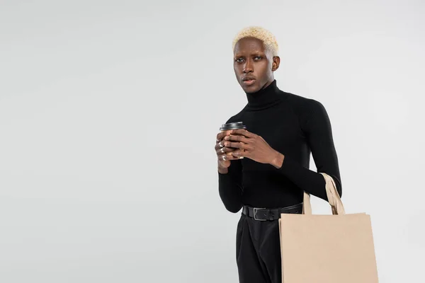 Blonde african american man standing with shopping bag and paper cup isolated on grey — Stock Photo