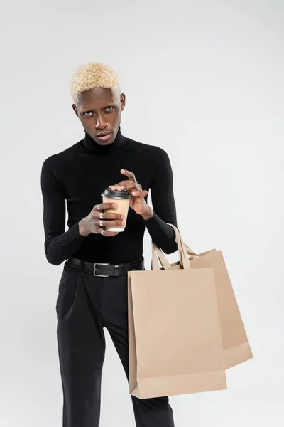 Hombre afroamericano elegante de pie con bolsas de compras y taza de papel aislado en gris - foto de stock