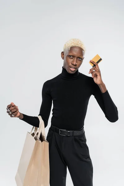 Loira afro-americana homem segurando sacos de compras e cartão de crédito isolado no cinza — Fotografia de Stock