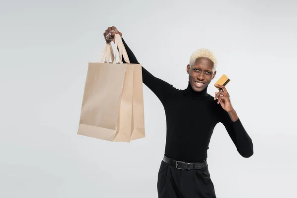 Alegre afroamericano hombre sosteniendo bolsas de compras y tarjeta de crédito aislado en gris - foto de stock