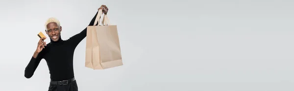 Happy african american man holding shopping bags and credit card isolated on grey, banner — Stock Photo