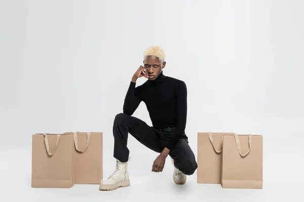 Longitud completa de hombre afroamericano rubia sentado cerca de bolsas de compras en gris - foto de stock