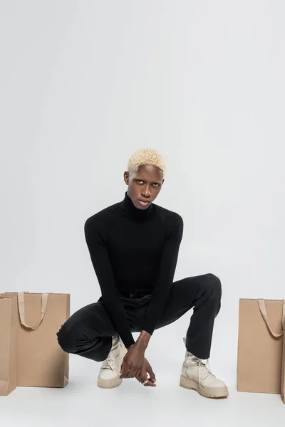 Pleine longueur de jeune homme afro-américain assis avec des sacs en papier sur gris — Photo de stock