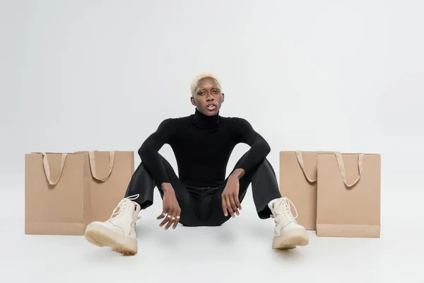 Longitud completa de hombre afroamericano rubia sentado cerca de bolsas de papel en gris - foto de stock