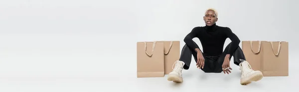 Full length of blonde african american man sitting near paper bags on grey, banner — Stock Photo