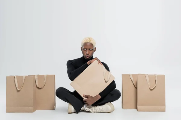 Comprimento total de homem americano africano loiro sentado com sacos de papel em cinza — Fotografia de Stock