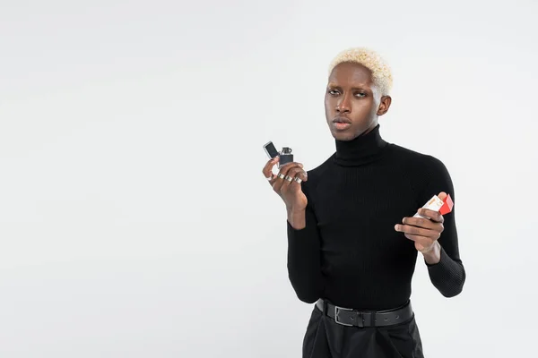 Jeune homme blond afro-américain tenant pack avec cigarettes et briquet isolé sur gris — Photo de stock