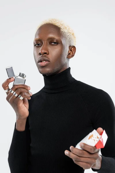 Blonde african american man holding pack with cigarettes and lighter isolated on grey — Stock Photo