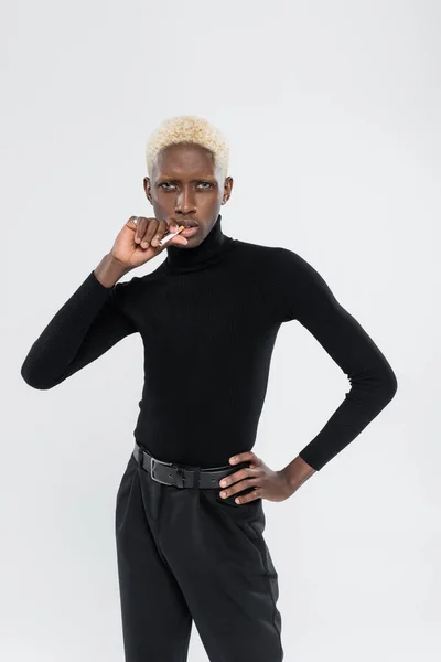 Young and blonde african american man holding cigarette and standing with hand on hip isolated on grey — Stock Photo