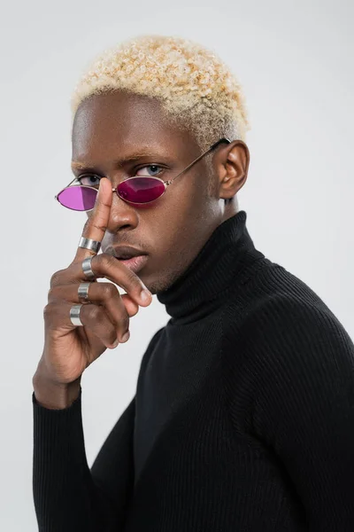 Biondo africano americano uomo in dolcevita regolazione occhiali da sole rosa isolato su grigio — Foto stock