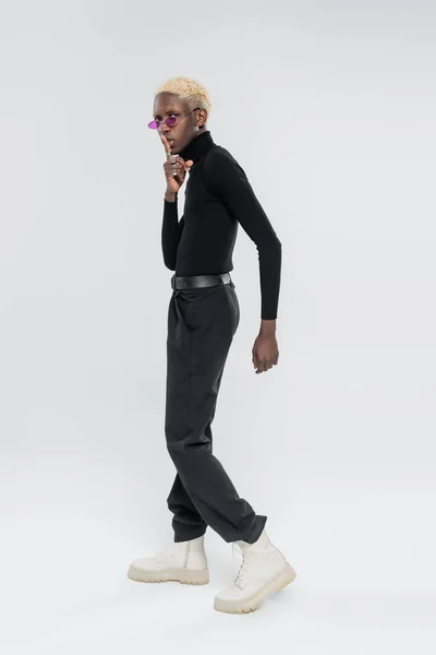 Full length of blonde african american man in turtleneck and pink sunglasses showing hush sign on grey — Stock Photo