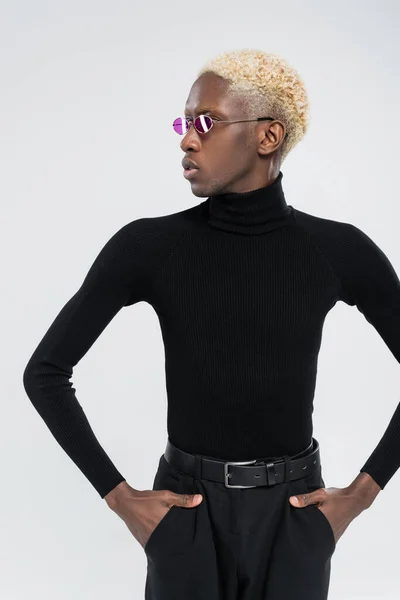 Blonde african american man in turtleneck and pink sunglasses posing with hands in pockets isolated on grey — Stock Photo